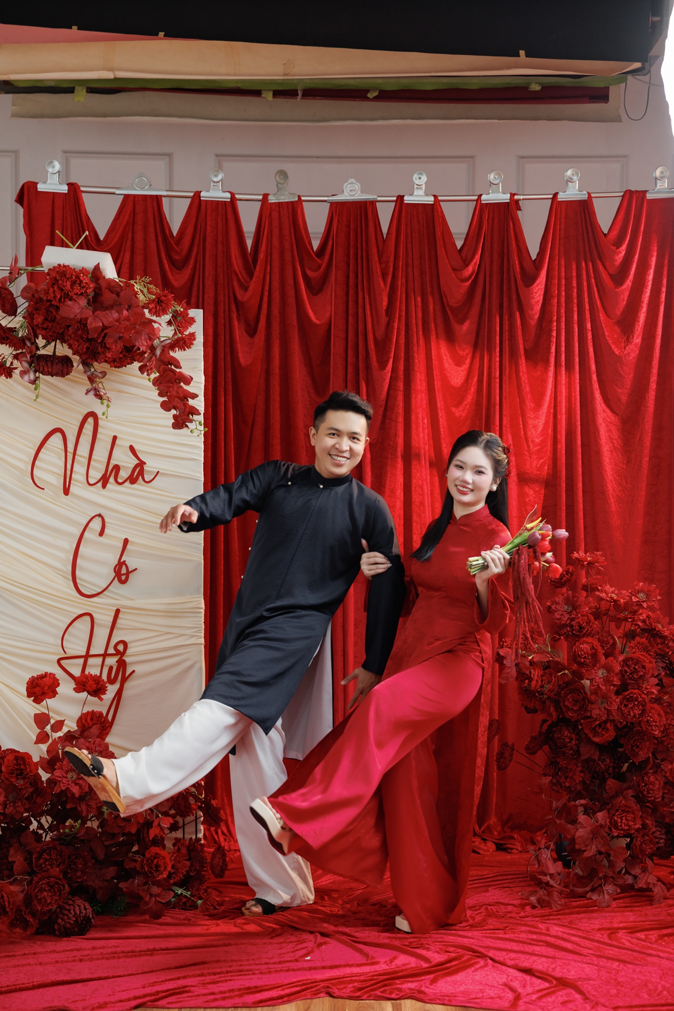 Bride in a white wedding dress and groom in a dark suit embracing and smiling, standing close in soft natural light with a blurred outdoor background and indistinct guests and decorations, warm joyful intimate atmosphere, no legible text in the image